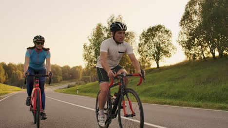 Un-Hombre-Y-Una-Mujer-En-Bicicleta-Recorren-Juntos-La-Carretera-Al-Atardecer-En-Cámara-Lenta.-La-Pareja-Viaja-En-Bicicleta.-Cascos-De-Ciclismo-Deportivos.